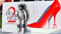A larger poster for "The Devil Wears Prada 2" on a red carpet is in the background with an oversized red heel, in the foreground is a spacesuit. 