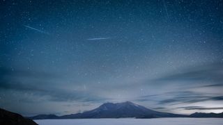 Several bright meteors streak through the sky over a white mountain on a blue lake