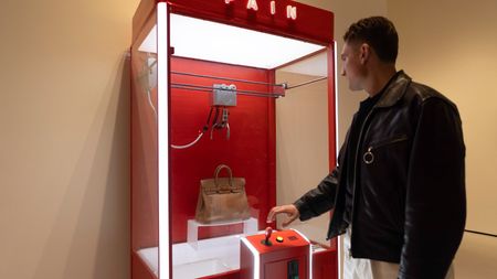 A man in a leather jacket stands before the red "PAIN" claw machine, reaching for the joystick to attempt to grab the designer handbag inside.