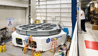A gray steel structure that simulates one of the massive 16.5-ton Giant Magellan Telescope primary mirror segments is installed onto a test cell. The GMT test cell and mirror simulator will be used to test the support structure and actuators that hold the massive telescope in place, including the software that controls the precise movements of the mirrors. 