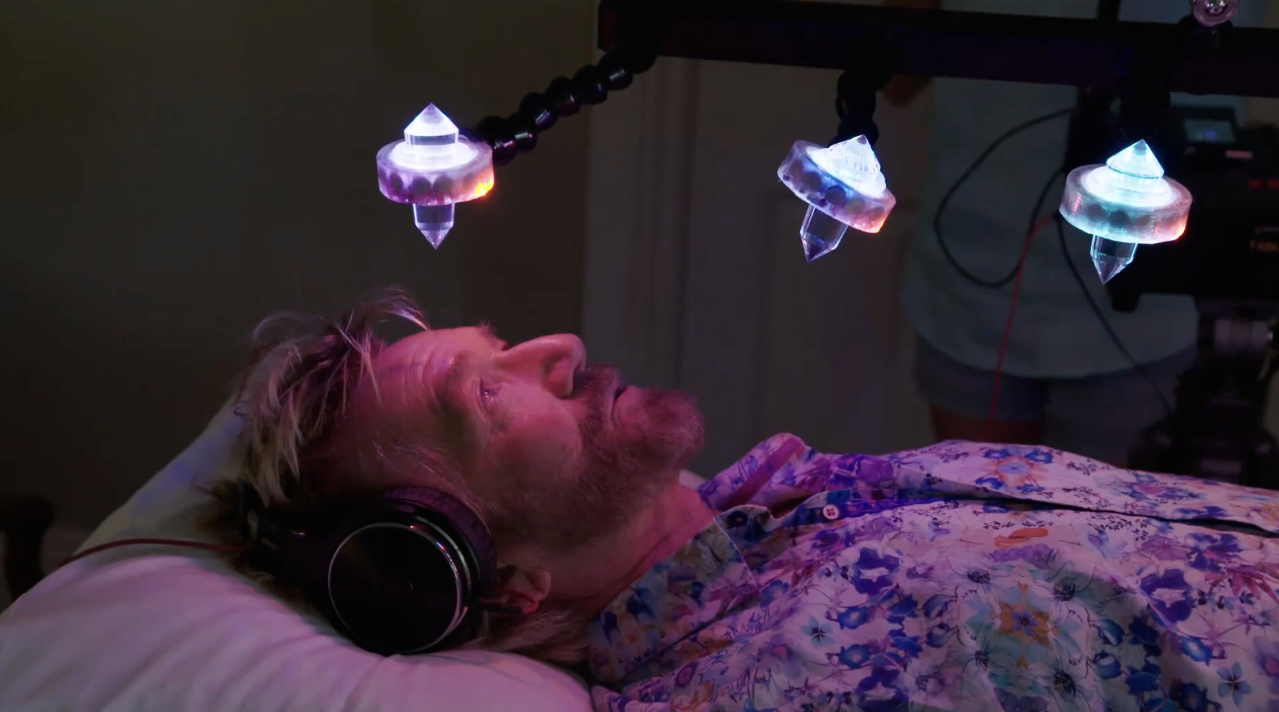 Noel lying down on his crystal bed, wearing headphones while crystals are light up and suspended above him.