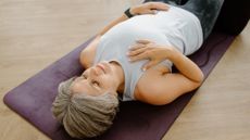 Woman lies on her back on a yoga mat with her eyes closed, one hand on her abdomen and one hand on her collarbone