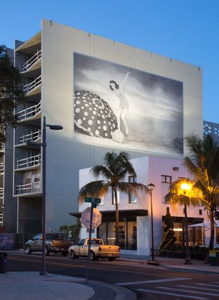 Miami street image, palm trees, road with parked trucks, street light, small square white building, windows, tall white panelled wall with black and white billboard poster of a girl on a beach, spotted parasol, clear blue dusk sky