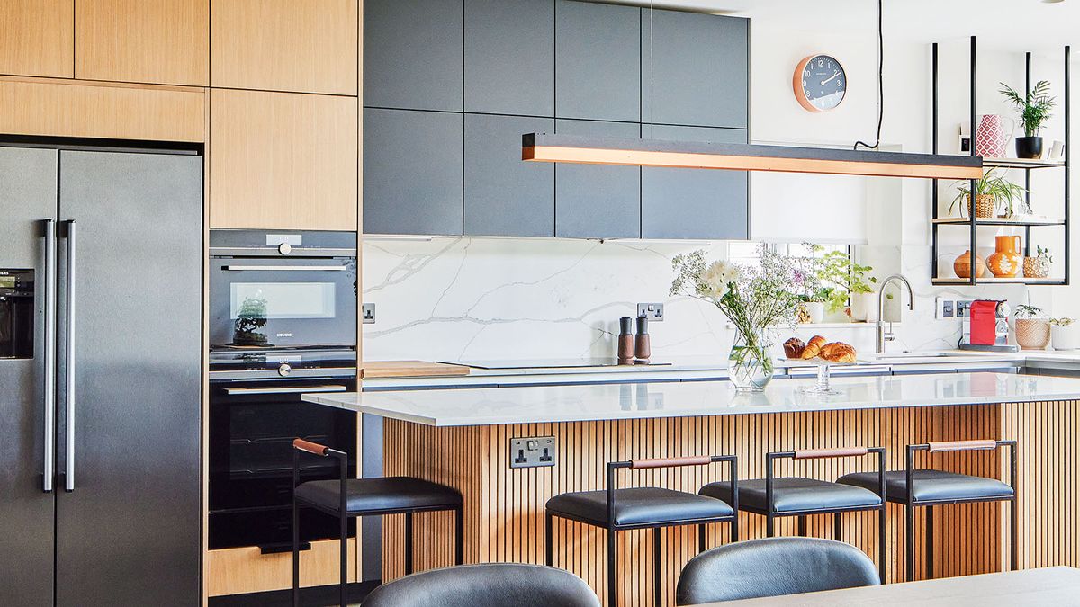 A newly fitted kitchen with a mixture of natural coloured plywood and dark cabinets. Thrre is wood panelling around the island and light surface. A long pendant light hangs over the kitchen island which has bar seating