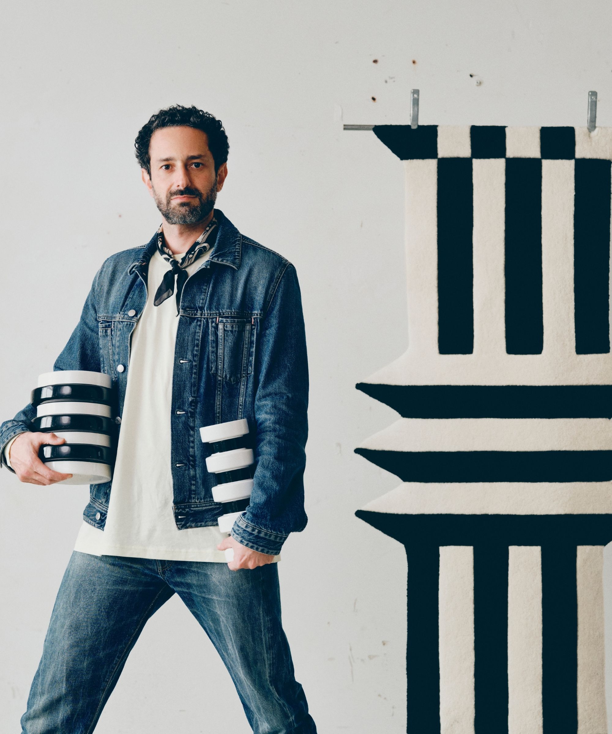 guy in denim jacket and jean holding black-and-white striped vase next to a black-and-white striped rug on the wall