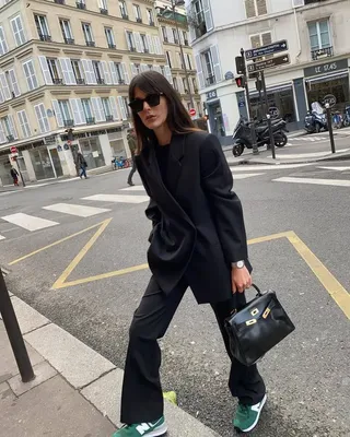 A woman wears a black blazer, black pants, black hermes bag, and green new balance sneakers.