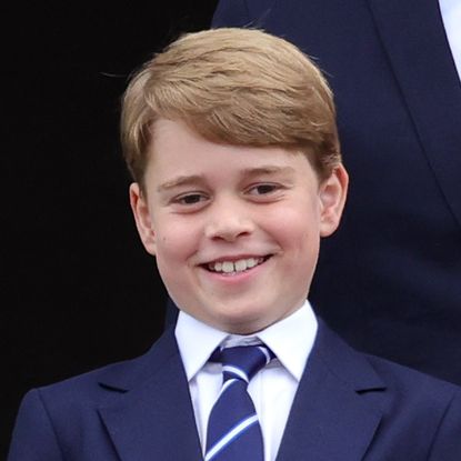 Prince George laughs while wearing a navy suit with a navy, red, and white striped tie, and Prince Harry wears a navy suit with his military medals attached