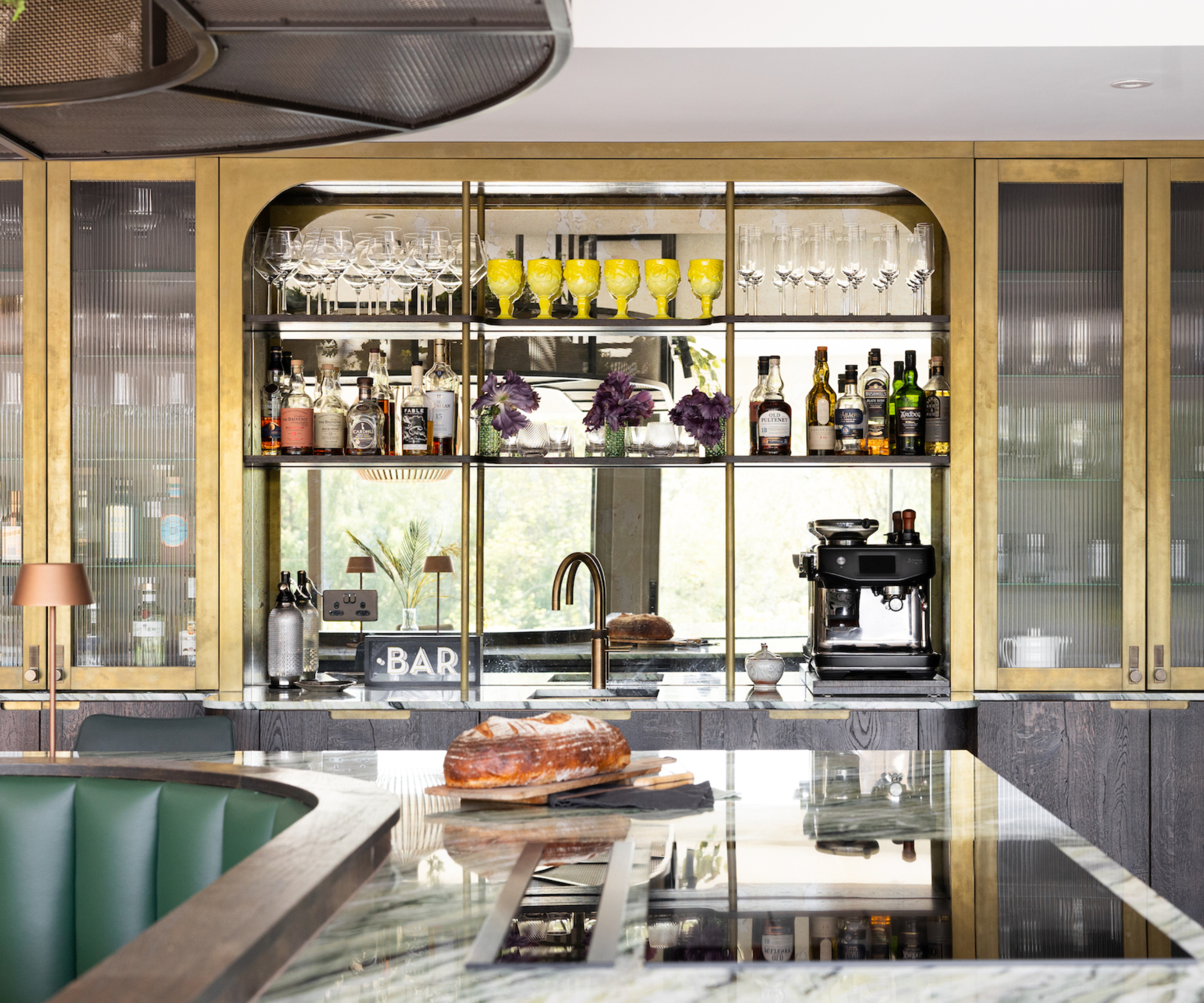 glass and gold kitchen cabinets around worktop and sink area with bar, coffee machine, sink and closed cabinets either side