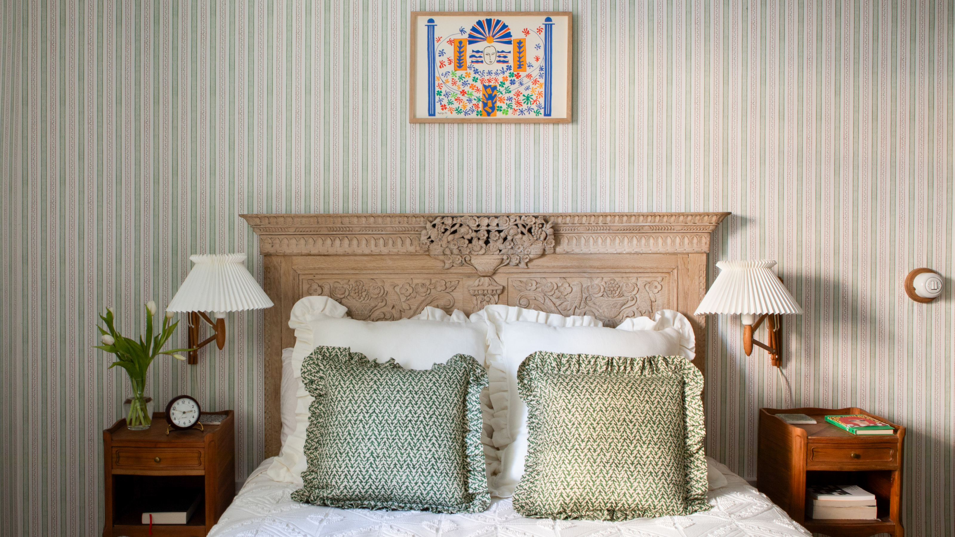 a classic bedroom with striped walls, an ornate bed frame, matching end tables, two sconces, and frilly pillows
