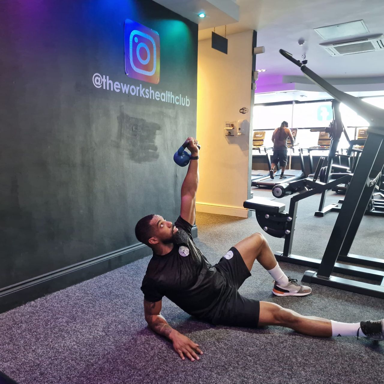 Renel Scarlett performs a kettlebell hip press inside The Works Health Club gym. He is seated on the floor, with his right leg straight and his left leg bent and planted on the ground. He is propped up on his right elbow, with his forearm on the ground, while his left arm is held straight up overhead and grasps a kettlebell. In the background we see treadmills.