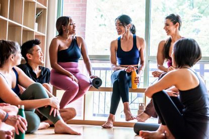 People talking in the reception area of a yoga studio. They are wearing sportswear and are in bare feet. The group are looking at a women with brown hair in a ponytail talking