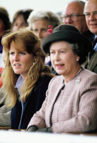 Sarah Ferguson has long red hair and bangs and sits next to Queen Elizabeth, who is wearing a checked coat and a hat