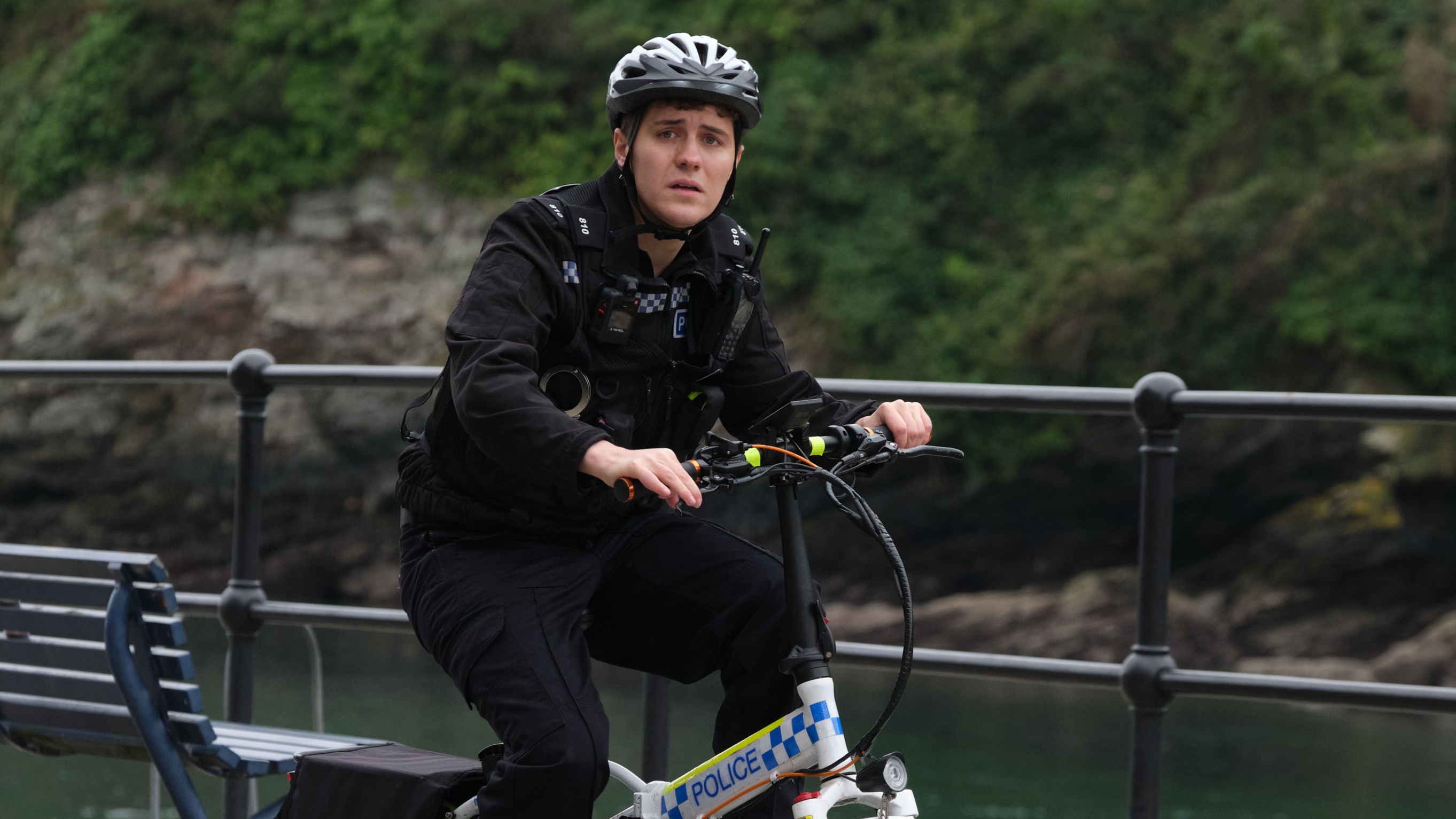 PC Kelby Hartford (Dylan Llewellyn) riding a police-branded electric bike along the waterfront. He is in his uniform and wearing a cycling helmet. He looks slightly worried. In the background there are metal railings and a bench, as well as trees and green foliage visible on the other side of the river.