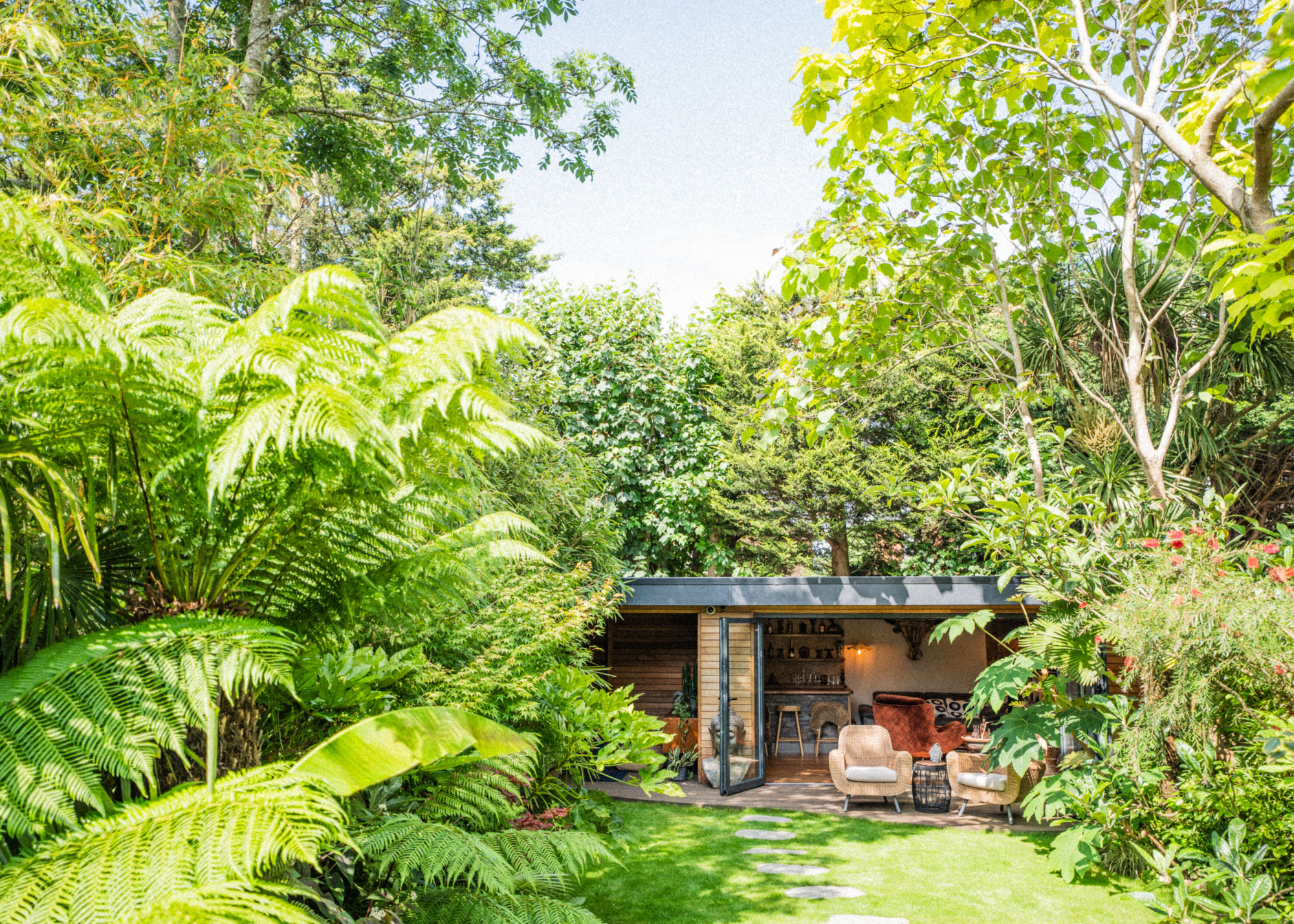 a garden room surrounded by large trees and greenery in a backyard. There is also outdoor furniture placed outside