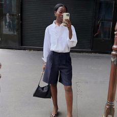 A woman wearing a white button up and blue long line shorts. 