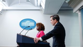 a person in a suit stands at a lectern under a sign reading "the white house"