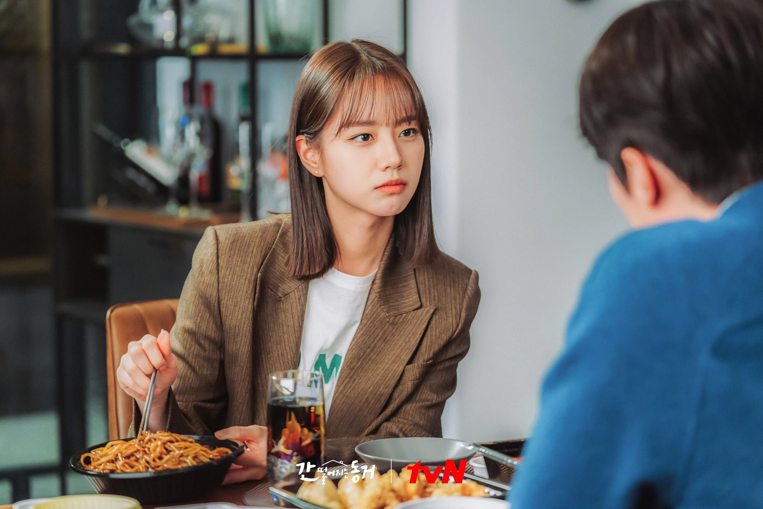A Korean woman (actress Lee Hye-ri) dips metal chopsticks into a noodle dish while sitting at a dining table with an unseen person, in the K-drama 'My Roommate Is a Gumiho.'