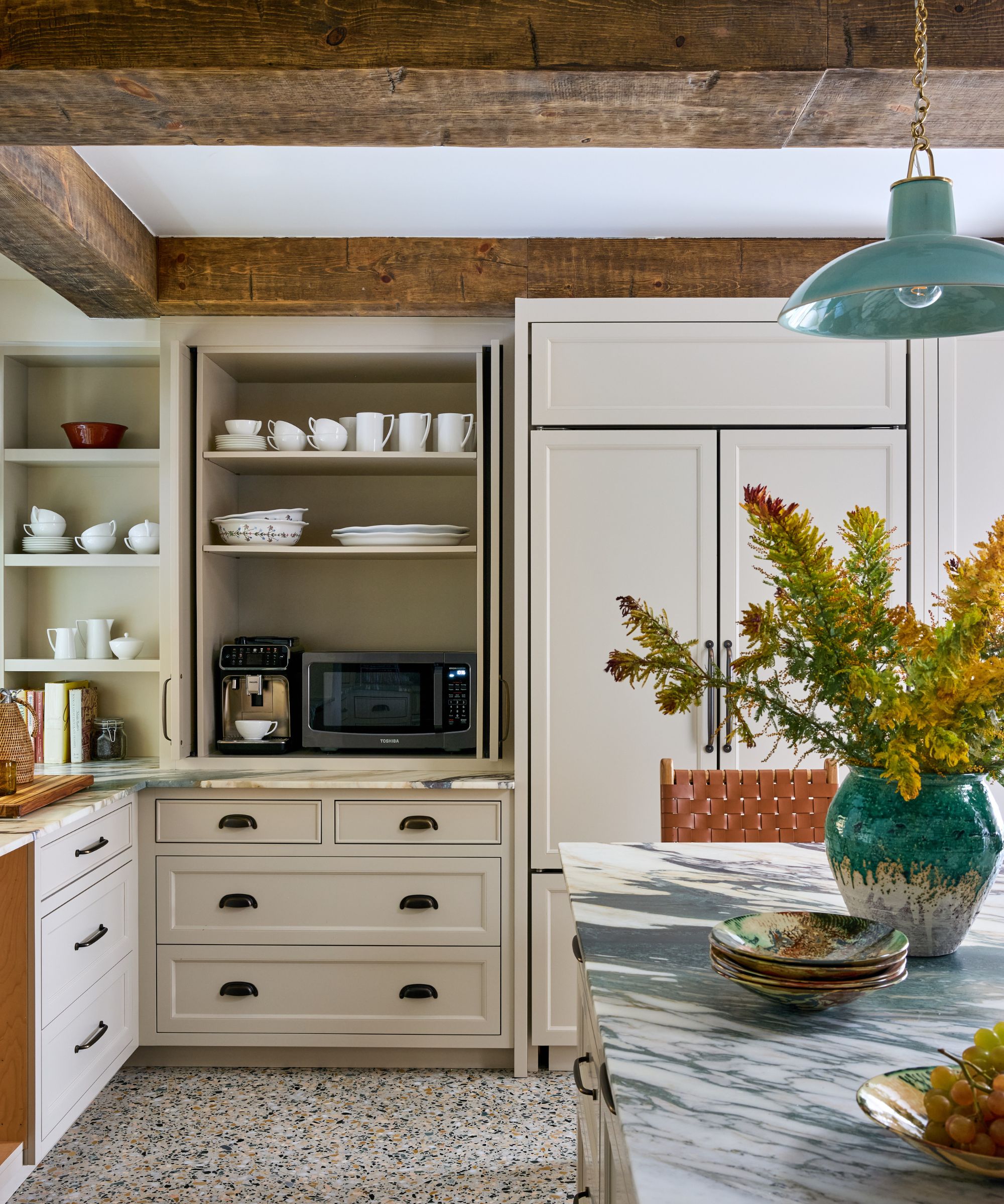 A kitchen with full-height neutral cabinets with open pantry storage, including a microwave and coffee machine. Wood ceiling beams and a marble kitchen island with a vase of yellow flowers.
