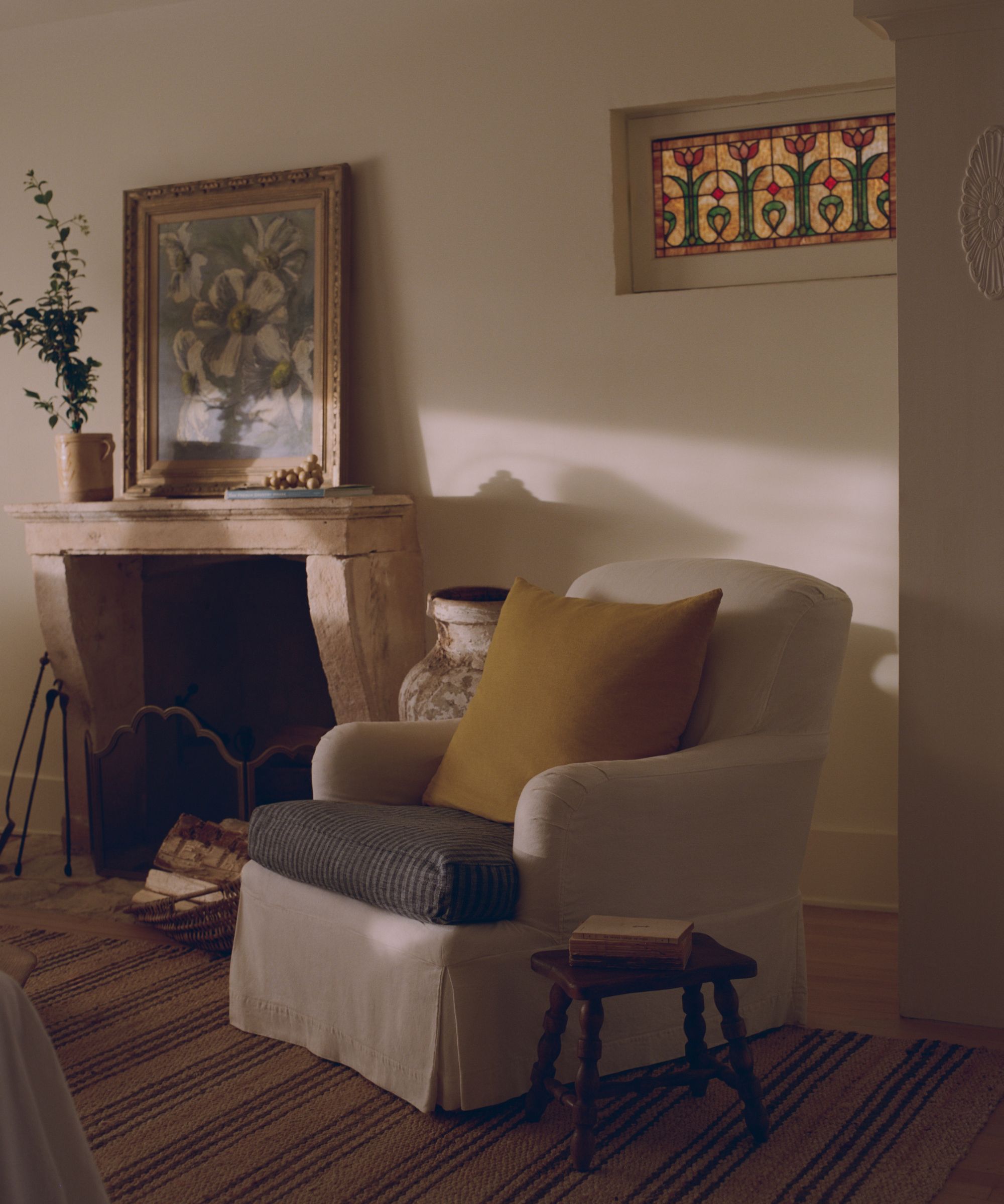 Warm, cozy living area featuring a white armchair with a mustard pillow, next to a stone fireplace and a stained glass window.