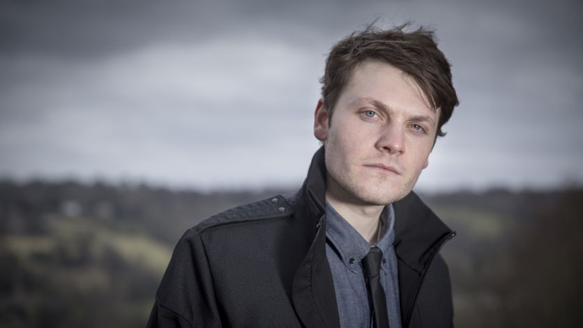 White man with dark hair, shirt and tie and black jacket, posing against a backdrop of a hillside and sky