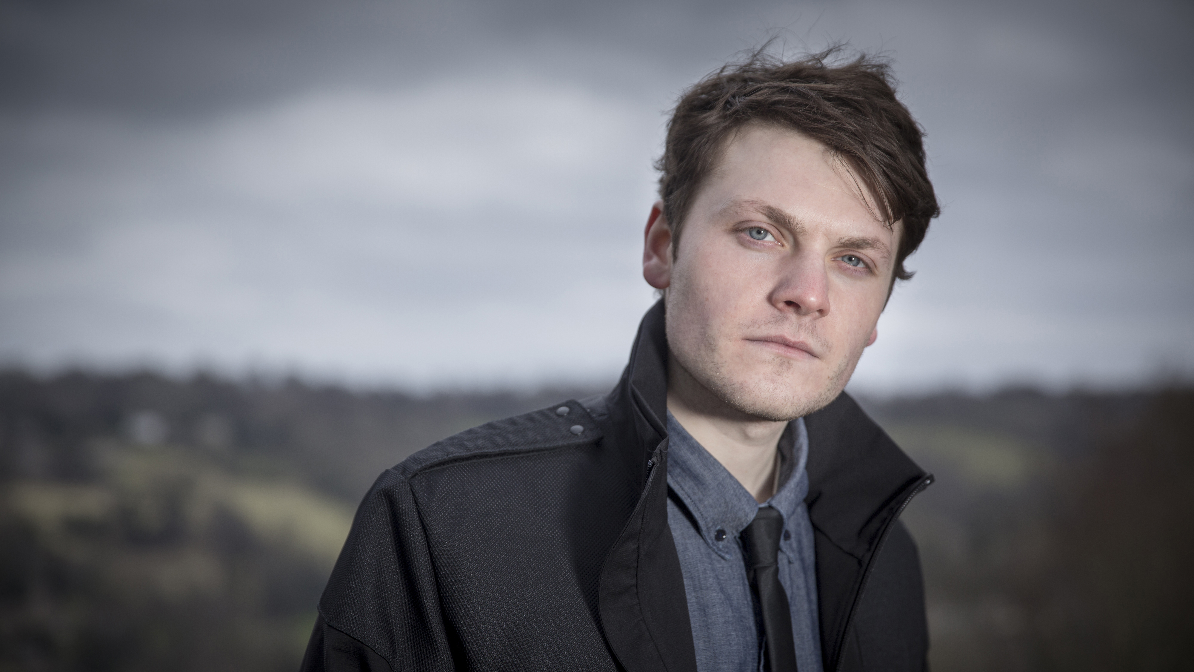 White man with dark hair, shirt and tie and black jacket, posing against a backdrop of a hillside and sky
