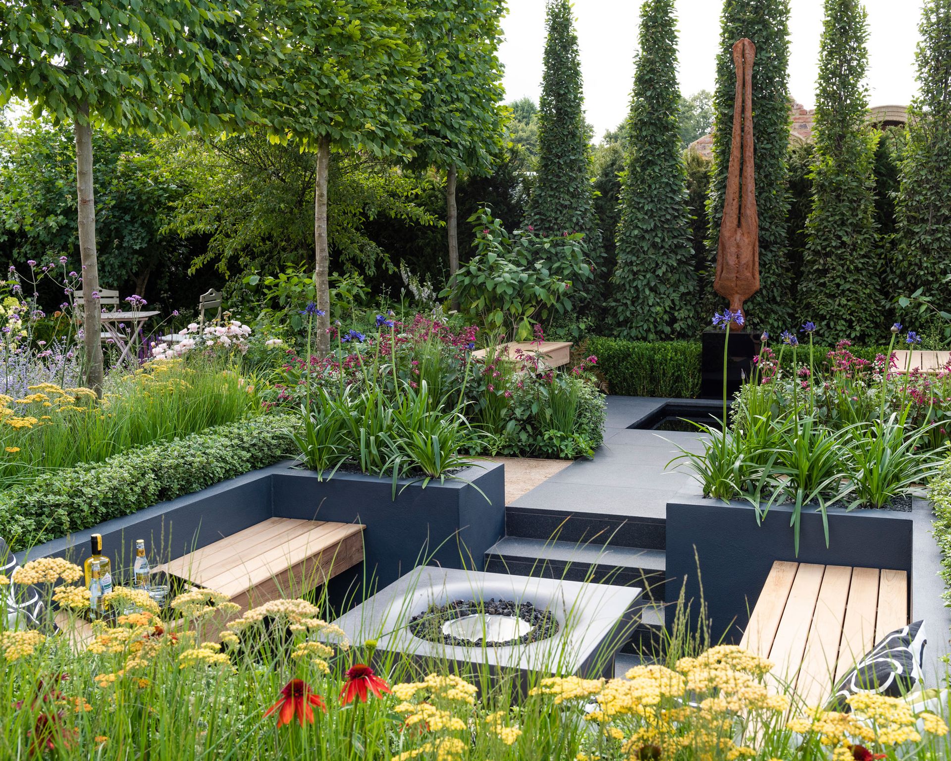 conversation pit enclosed by raised beds planted with agapanthus and topiary, with a fire table and wooden benches