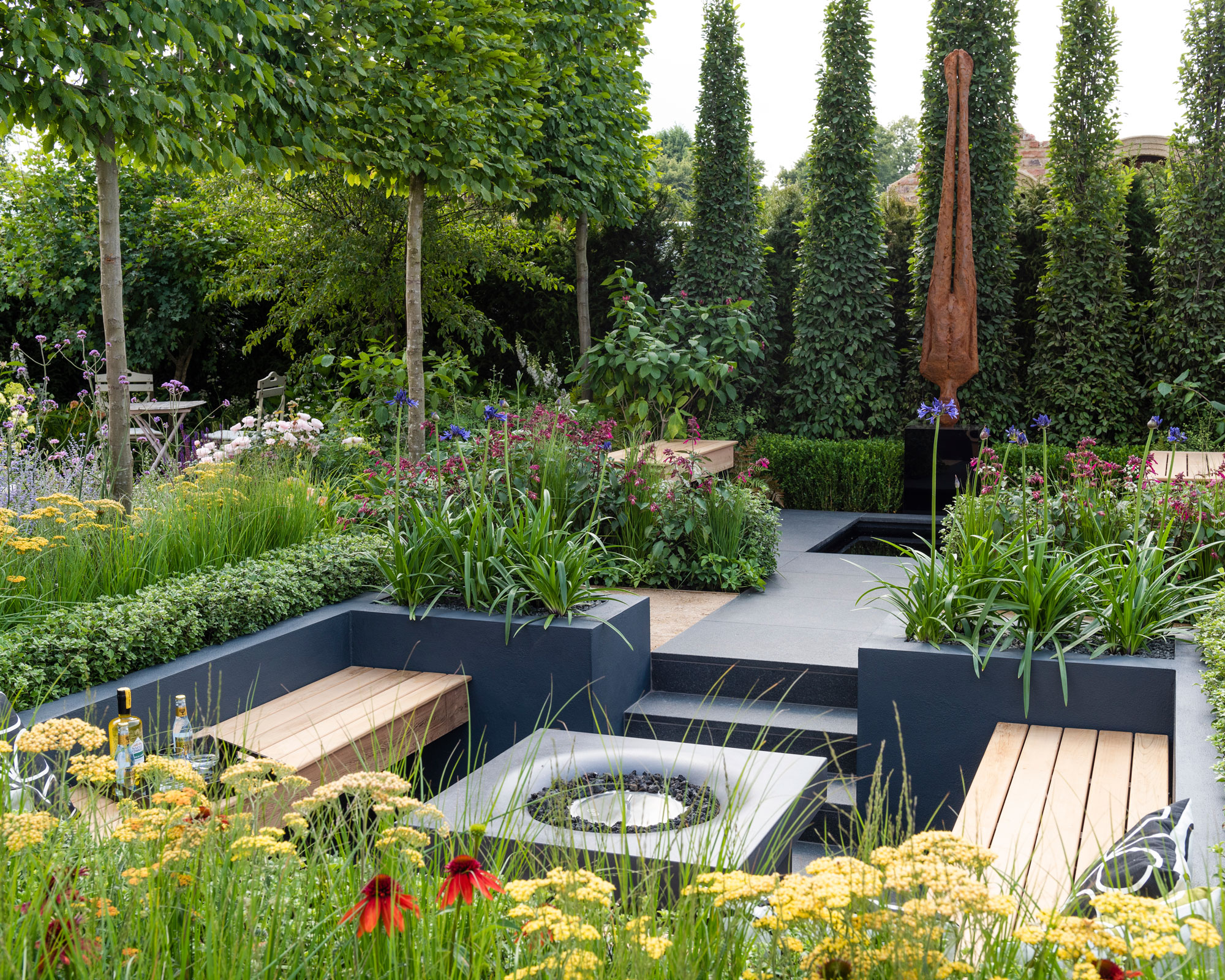 conversation pit enclosed by raised beds planted with agapanthus and topiary, with a fire table and wooden benches