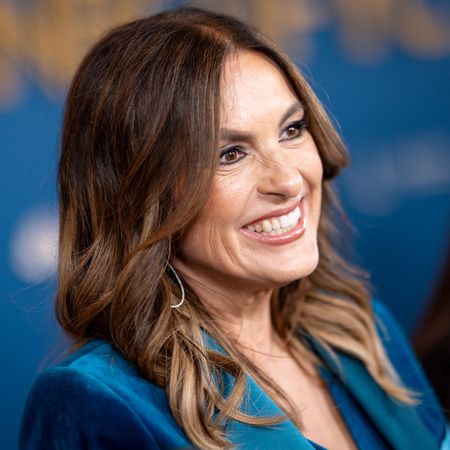 a close-up photo of a white woman with brown hair smiling on a red carpet in front of a blue backdrop
