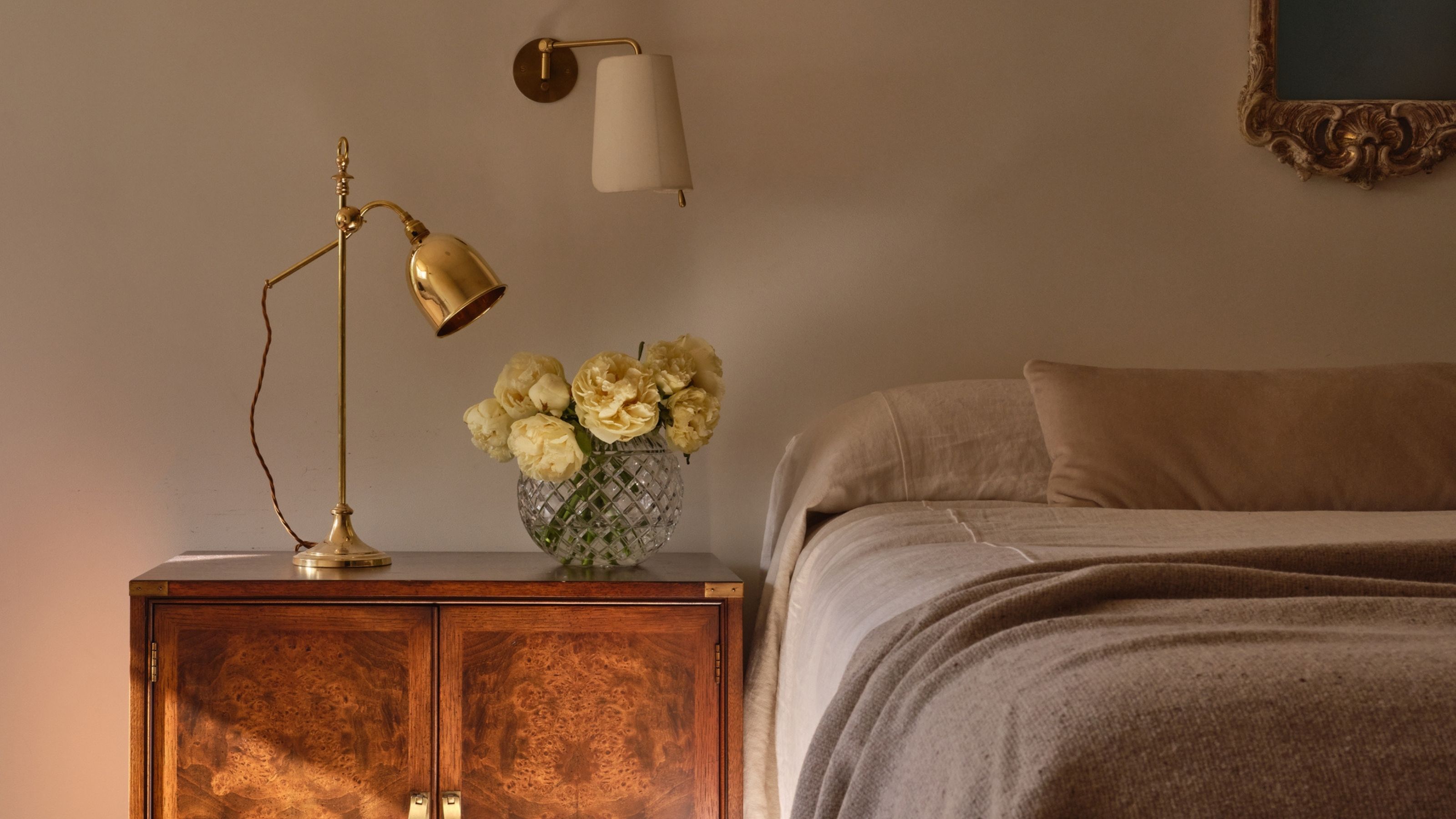 Image of a neutral bedroom with taupe walls and minimalist taupe bedding. There is a vintage-looking wooden bedside table with a gold lamp and a vase of yellow roses.