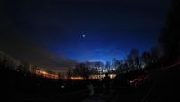 A bright, star-like planet shines in the evening sky at sunset above a treeline.