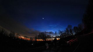 A bright, star-like planet shines in the evening sky at sunset above a treeline.