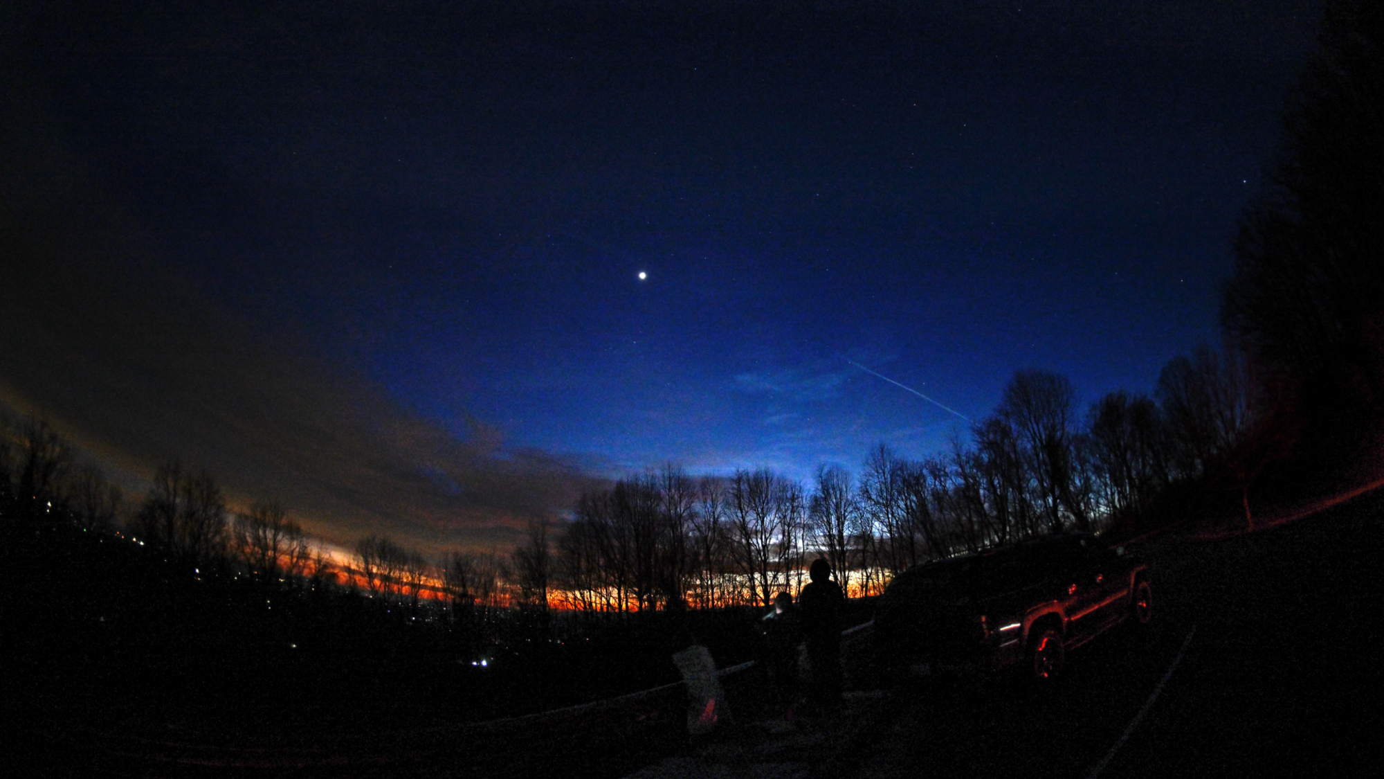 A bright, star-like planet shines in the evening sky at sunset above a treeline.
