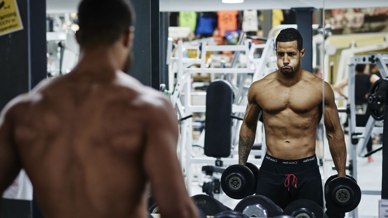 Determined muscular guy looking in the mirror in the gym before his dumbbell workout commences