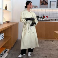 Mimi Nguyen in a New York City store wearing a low bun, black leather gloves, green suede clutch bag, belted trench coat, black pants, and Adidas Tokyo sneakers