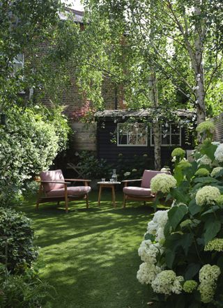 A garden with two red chairs and a wooden table on a small lawn surrounded by foliage and a hydrangea bush