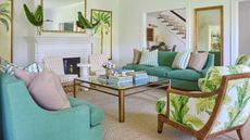 warm vs cool, green and neutral living room with leafy green upholstered armchair, two green couches, coir carpet, glass and metal coffee table, mirrors, patterned couch pillows, staircase in background