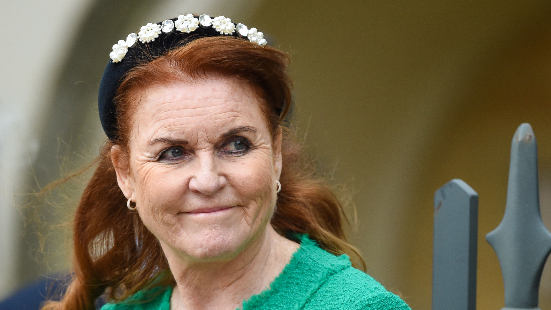 Sarah Ferguson wearing a green jacket and black headband looking to the side and smirking