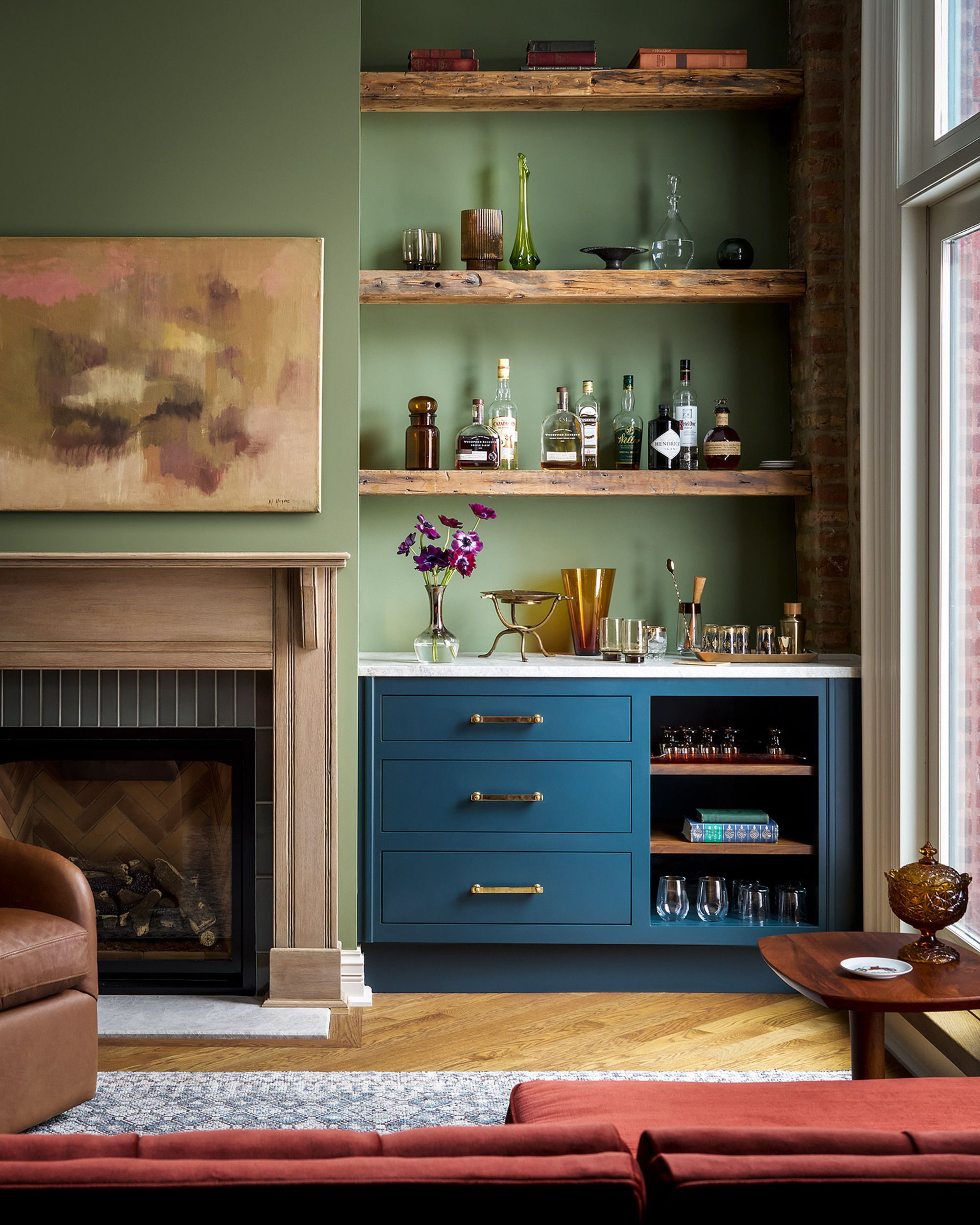 Teal blue cabinet in a living room next to a wooden fireplace. The walls are a mint green with matching wooden shelving. The room used earthy tones with golden fixtures 