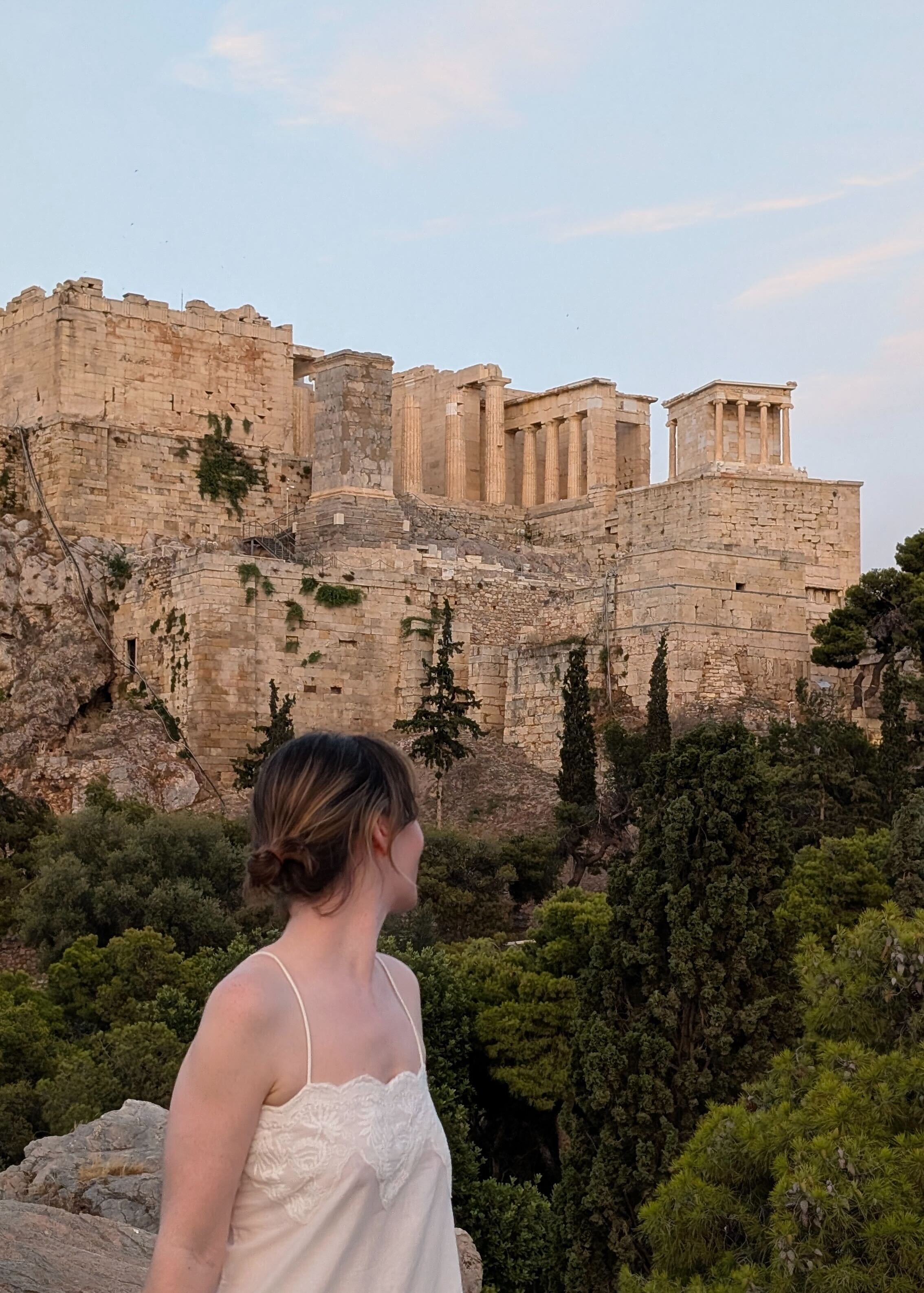 A young woman dressed in a white linen blouse dress and with her hair gathered in a low bun stares at ancient ruins immersed in green while facing the camera with her neck.