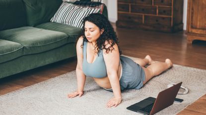 Woman practicing Pilates swan variation exercise in living room in front of laptop