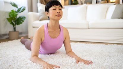 woman in a lilac vest performing sphinx pose facing the camera on a white fluffy living room rug with a white sofa behind her.