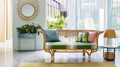 sofa trends to avoid. Belladonna rattan sofa with green seat cushion on screed floor in front of window with light blue sheer curtains, basket side table and rattan mirror on wall.