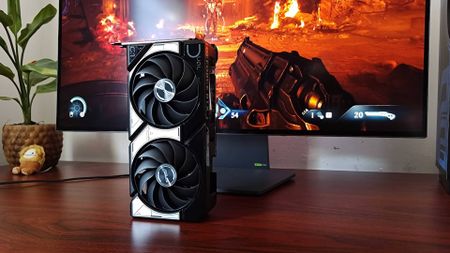 RTX 5060 graphics card standing upright with DOOM: The Dark Ages gameplay on monitor in backdrop.