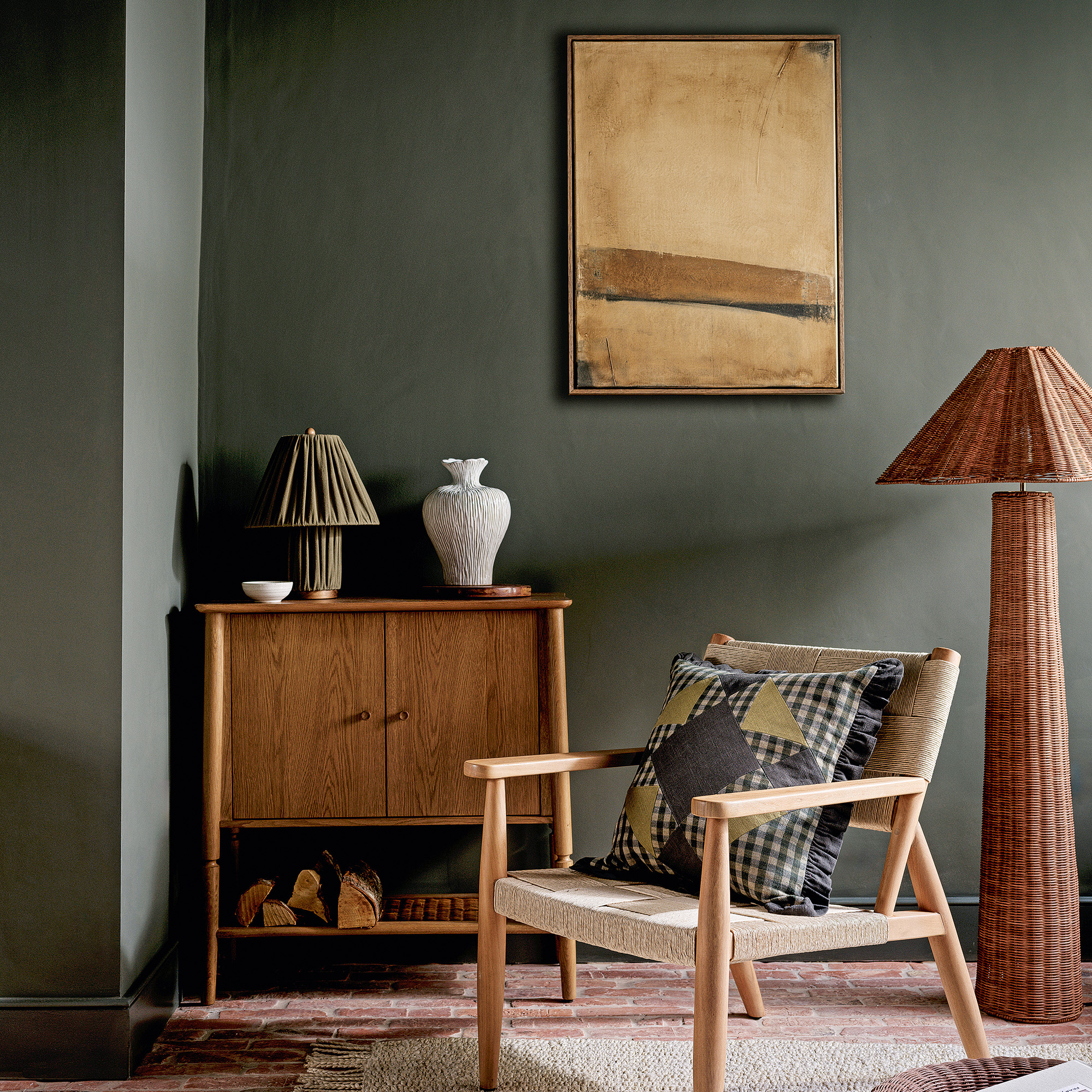 living room with moody green matt walls wooden side board a wooden amrchair with gingham patchwork cushion and a rattan floor standing lamp neutral toned abstract artwork in the centre of the room