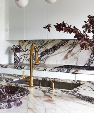 white gloss kitchen finished with bold veined marble countertop and backsplash with a large brass faucet