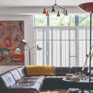 A living room with a large brown leather modular sofa, a colourful ceiling light and a floor lamp by a painting