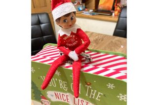 The Elf on the Shelf pictured sitting on a suitcase that says 'You're on the nice list'