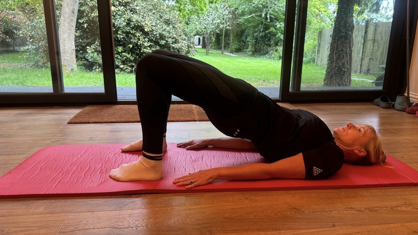 Sarah Haselwood performs a glute bridge at home on an exercise mat. She is on her back, with her shoulders on the mat, knees bent, feet on the floor and hips elevated, so that her body forms a straight line from her knees to her chest.