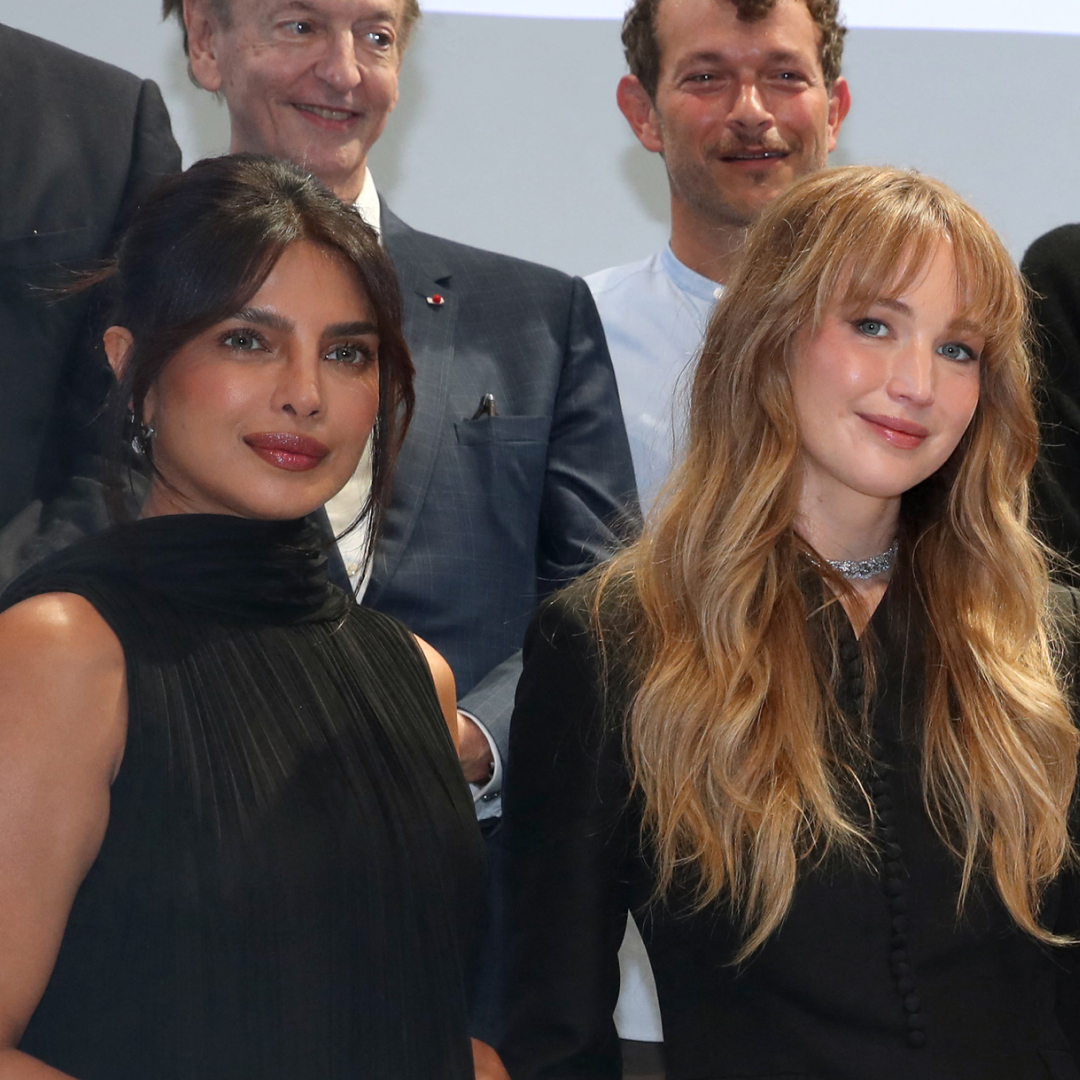 Jennifer Lawrence has long blonde hair and is wearing a black Dior suit, Priyanka Chopra wears her black hair tied back and wears a black Dior dress with a white tiered skirt