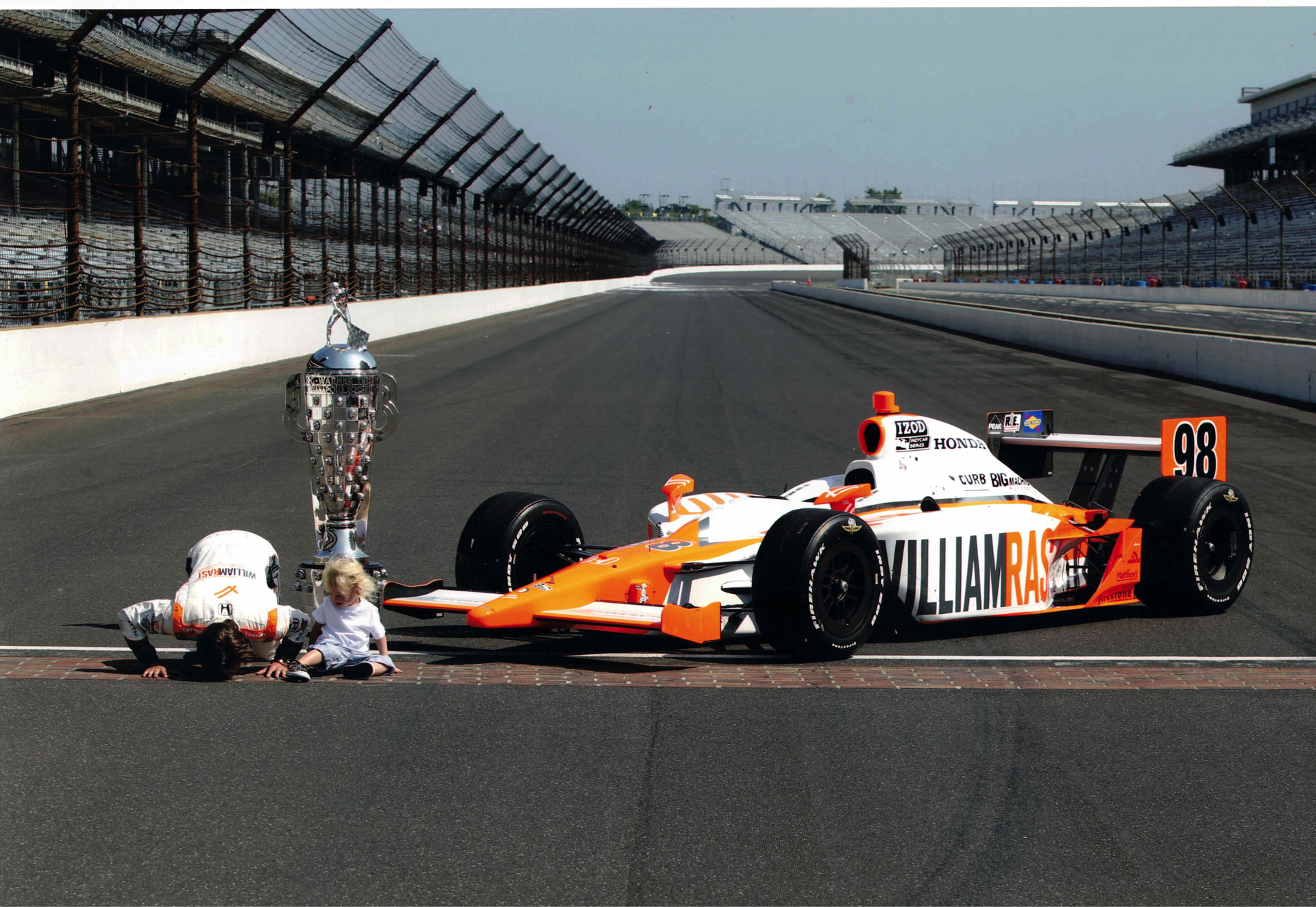 a man (dan wheldon) bows to the camera as his young son (sebastian wheldon) sits next to him on a racetrack; the pair are to the left of a large silver trophy and an orange and black race car